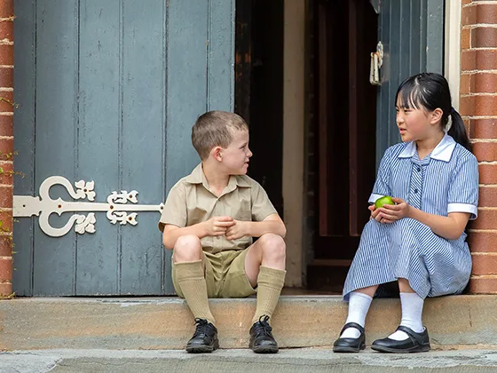 Two students, a boy and a girl, sit on a stoop next to an open door. The girl is holding an apple.